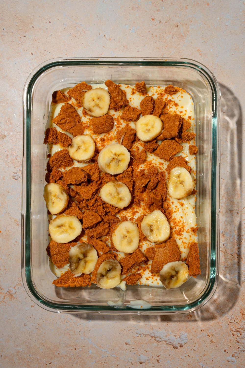 A glass baking dish with a layer of Biscoff banana pudding including pudding, Biscoff cookie pieces, and sliced bananas. The dish is on a tan counter.