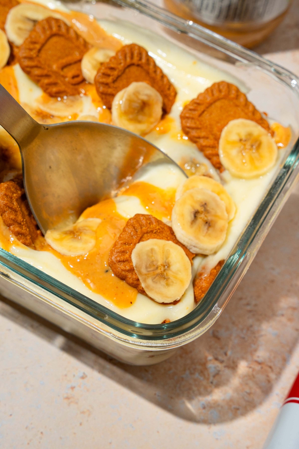 A glass baking dish with Biscoff banana pudding topped with Biscoff cookie pieces, sliced bananas, and caramel drizzle. A spoon is taking a bite of pudding.