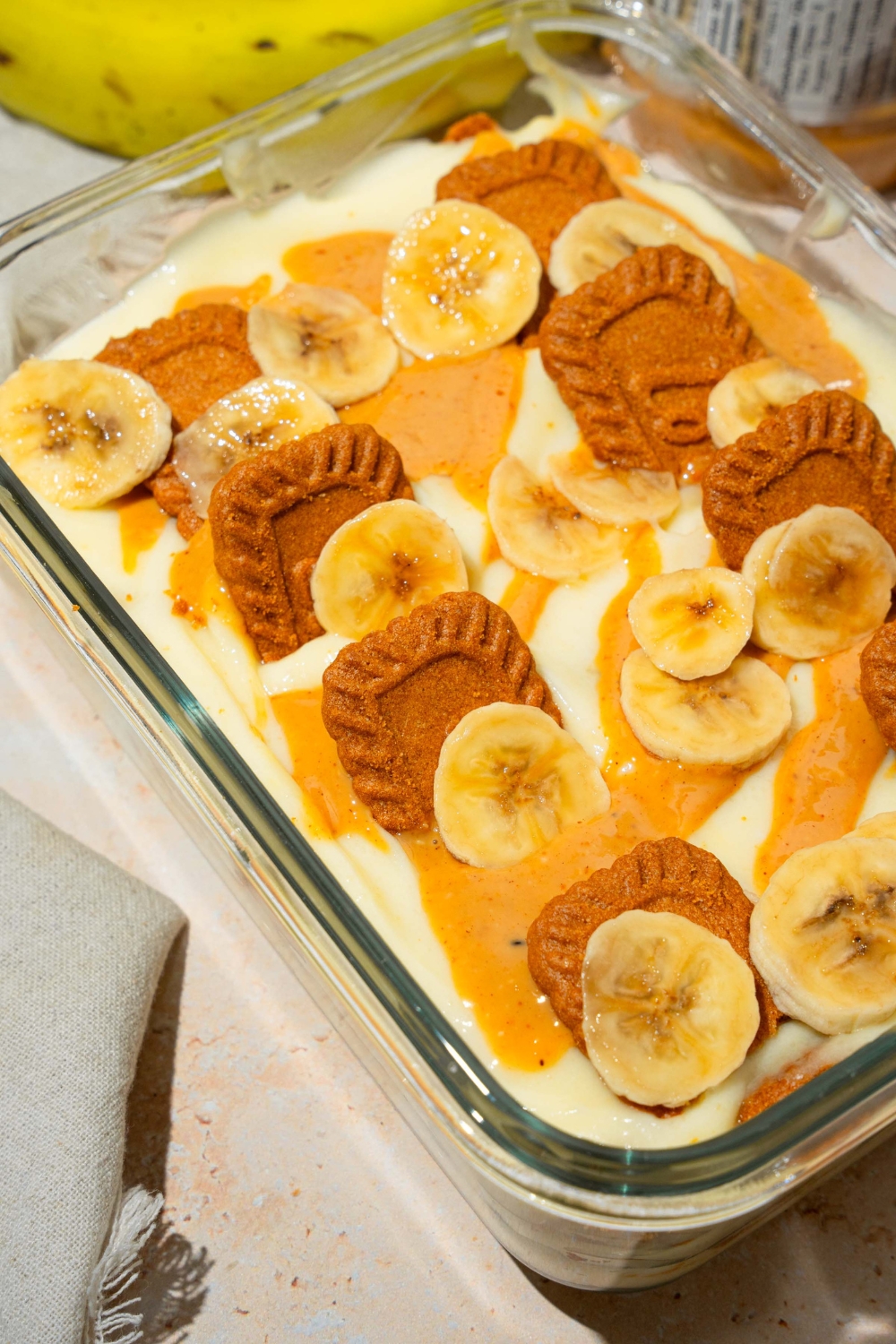 A glass baking dish with Biscoff banana pudding topped with Biscoff cookie pieces, sliced bananas, and caramel drizzle. The dish is on a tan counter with bananas.