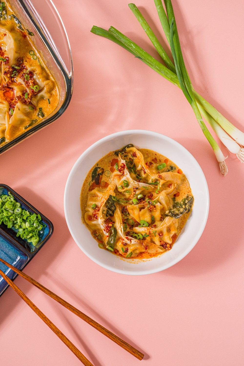 A white bowl with Trader Joe's chili crisp dumpling bake garnished with sliced green onion and Trader Joe's chili onion crunch. A pair of chopsticks is taking a dumpling from the bowl. The bowl is on a pink counter with a pair of chopsticks and green onions.