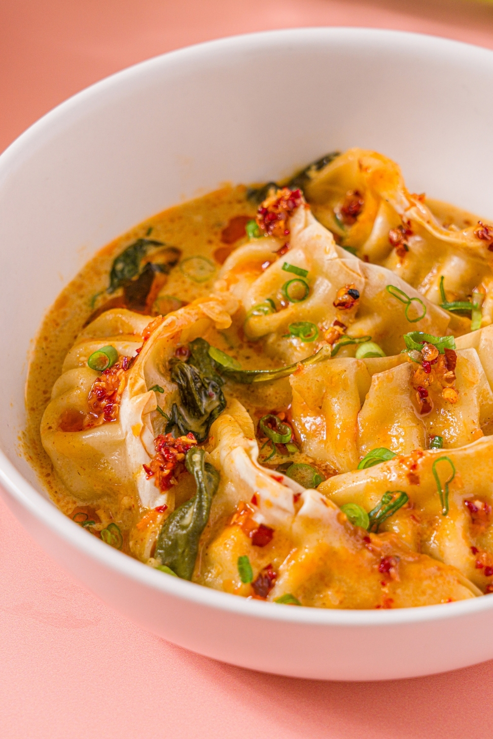 A white bowl with Trader Joe's chili crisp dumpling bake garnished with sliced green onion and Trader Joe's chili onion crunch. A pair of chopsticks is taking a dumpling from the bowl. The bowl is on a pink counter.