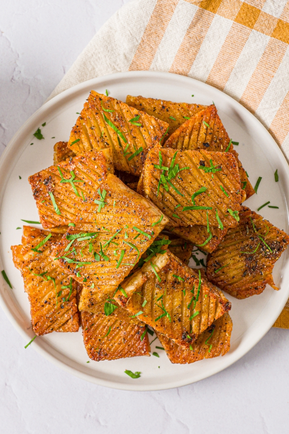 A white plate with seasoned baked accordion potatoes garnished with fresh parsley. The plate is on a white counter with a yellow checkered napkin.