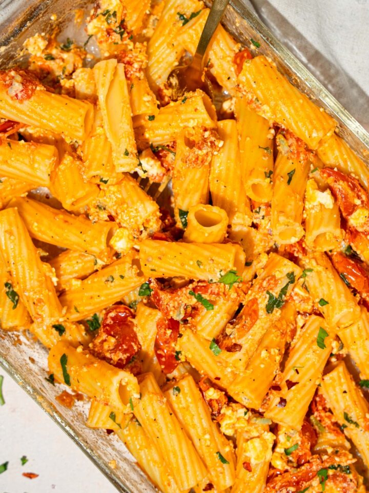 A glass baking dish with baked feta pasta garnished with fresh parsley and crushed red pepper. A fork is in the pasta. The dish is on a tan counter with a white cloth napkin.