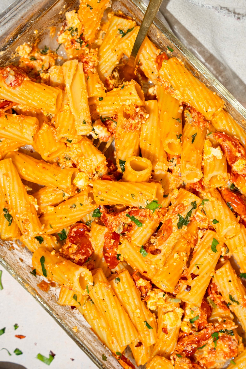 A glass baking dish with baked feta pasta garnished with fresh parsley and crushed red pepper. A fork is in the pasta. The dish is on a tan counter with a white cloth napkin.