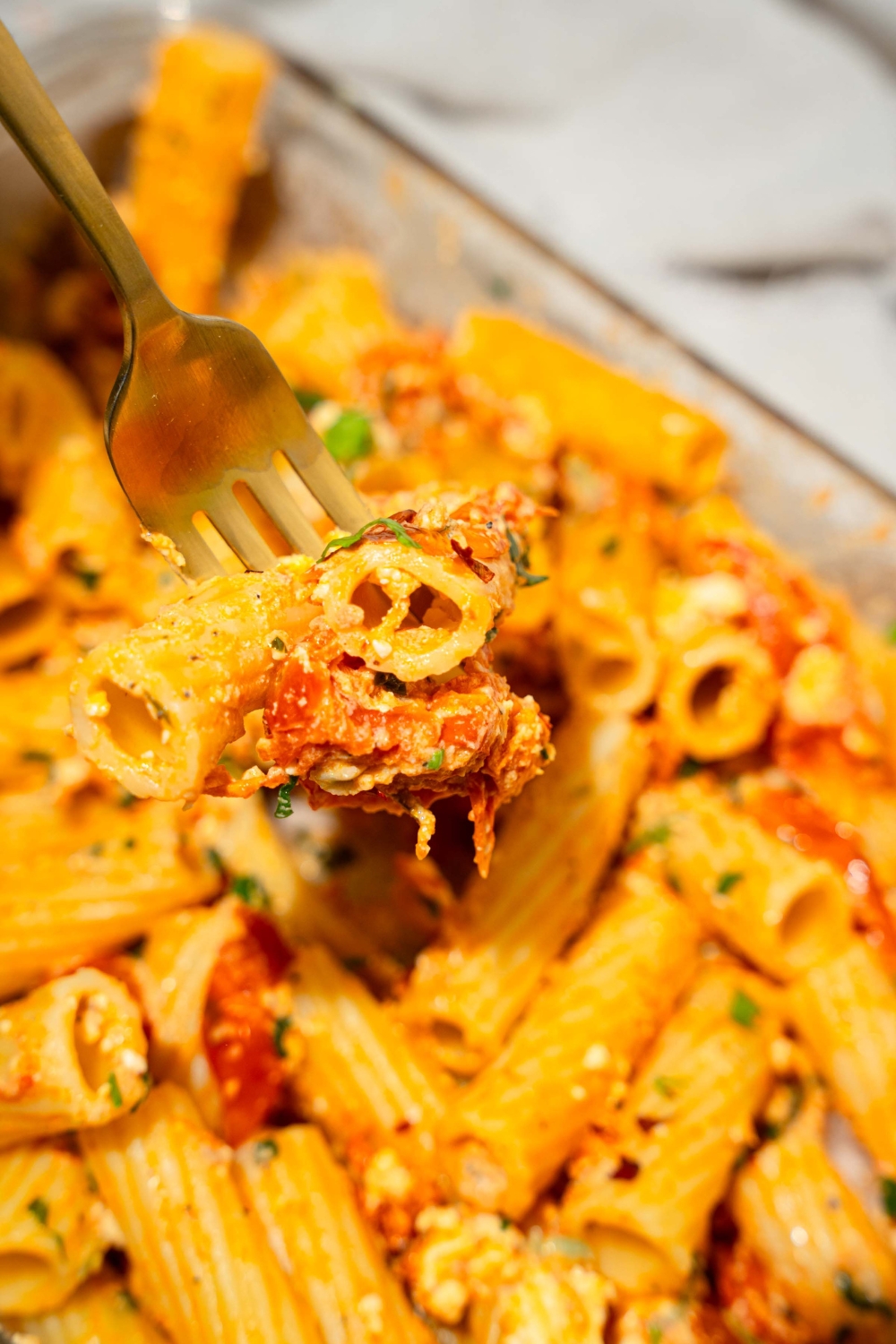 A glass baking dish with baked feta pasta garnished with fresh parsley and crushed red pepper. A fork is taking a bite of the pasta.
