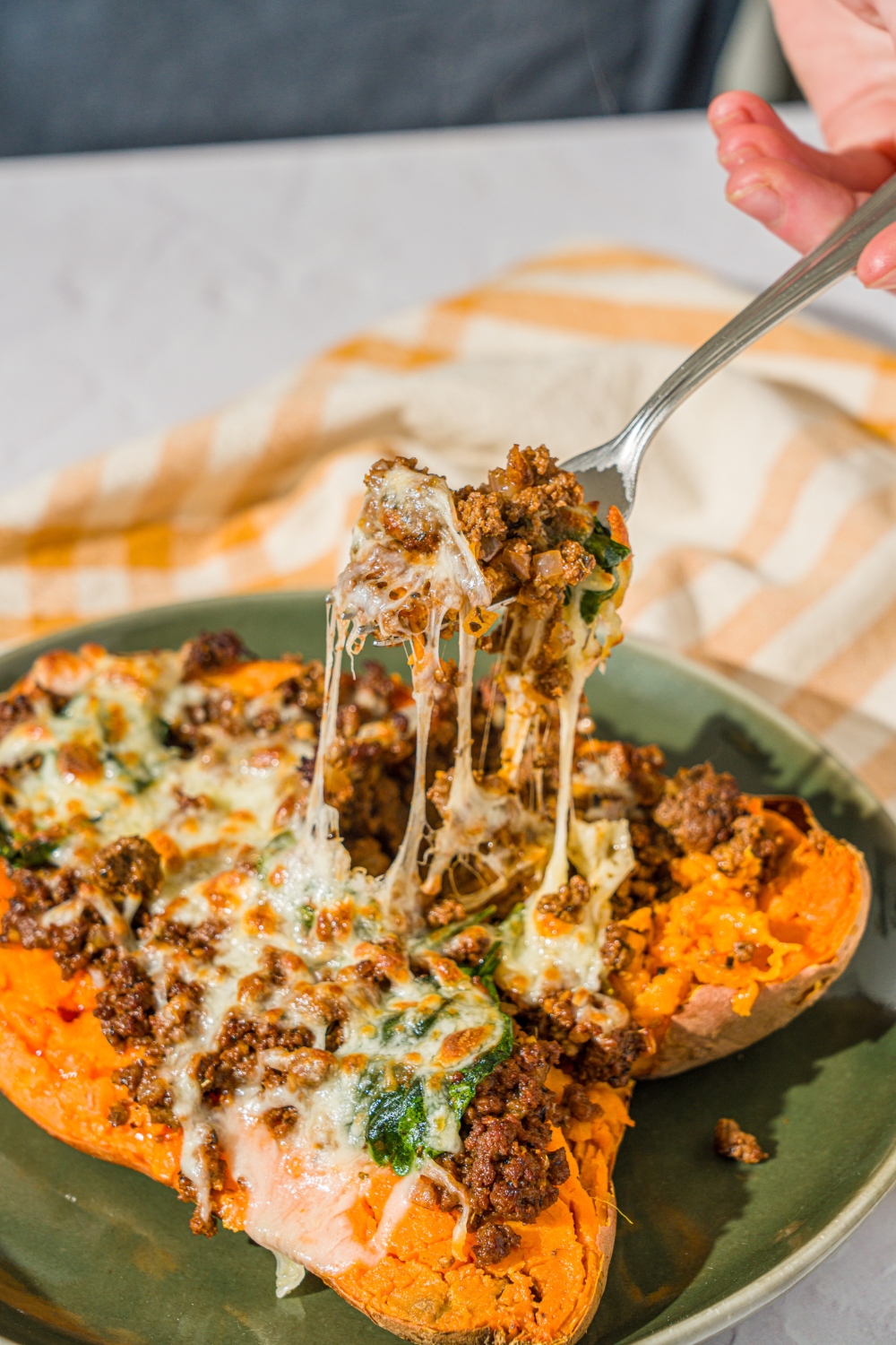 A green plate with a loaded sweet potato sliced open and stuffed with a beef mixture and topped with melted cheese. There is a fork taking a bite from the potato. The plate is on a white counter with a yellow checkered napkin.