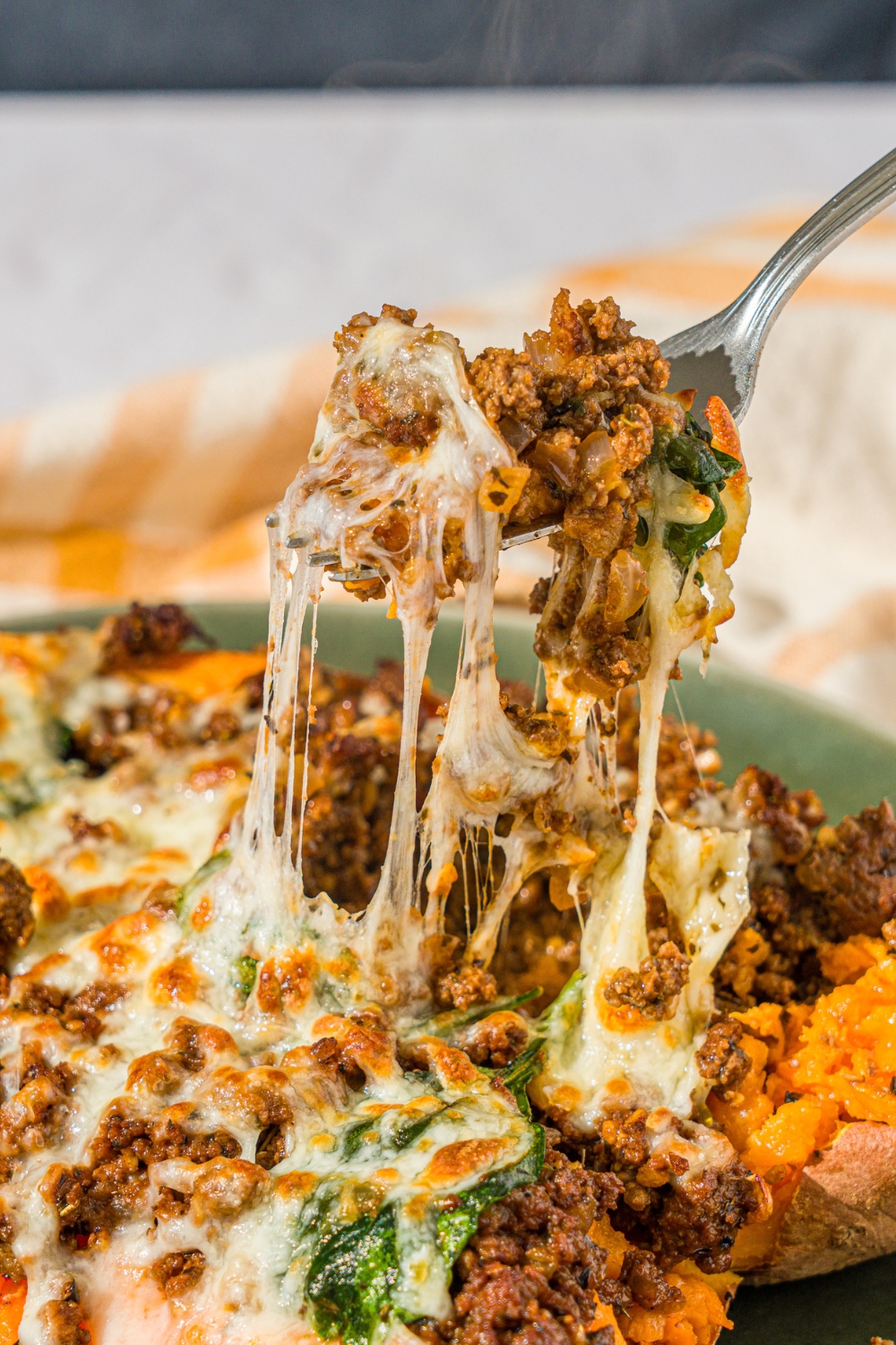 A green plate with a loaded sweet potato sliced open and stuffed with a beef mixture and topped with melted cheese. There is a fork taking a bite from the potato. The plate is on a white counter with a yellow checkered napkin.