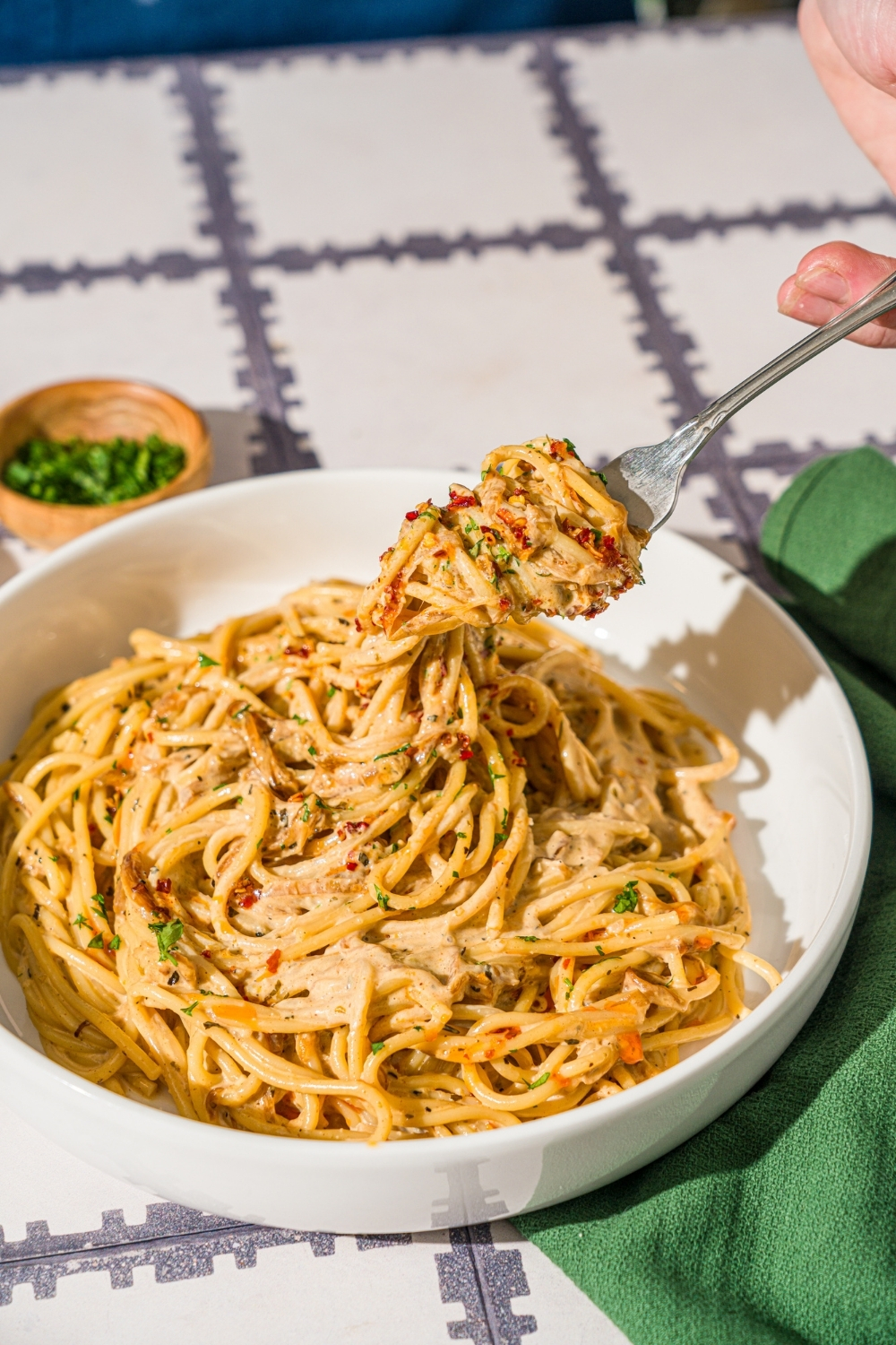 A white bowl with caramelized onion pasta garnished with chili crisp onion and parsley. A fork is twirling a bite of pasta from the bowl. The bowl is on a tiled counter with a green cloth napkin and bowls of garnishes.