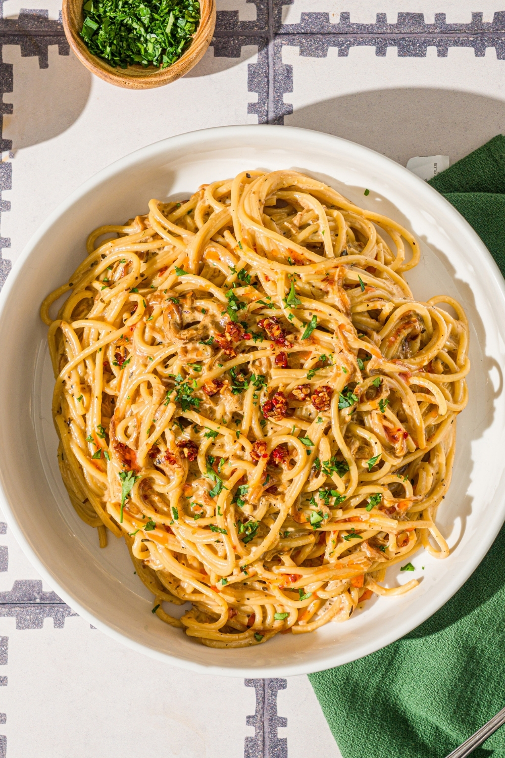 A white bowl with caramelized onion pasta garnished with chili crisp onion and parsley. The bowl is on a tiled counter with a green cloth napkin and bowls of garnishes.