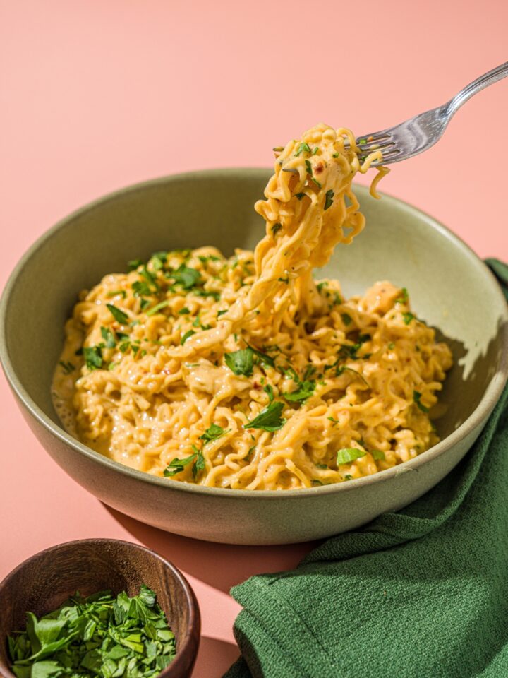 A green bowl with cheesy maggi noodles garnished with fresh cilantro. There is a fork taking a bite of noodles from the bowl. The bowl is on a pink counter with a green cloth napkin and small bowl of garnishes.