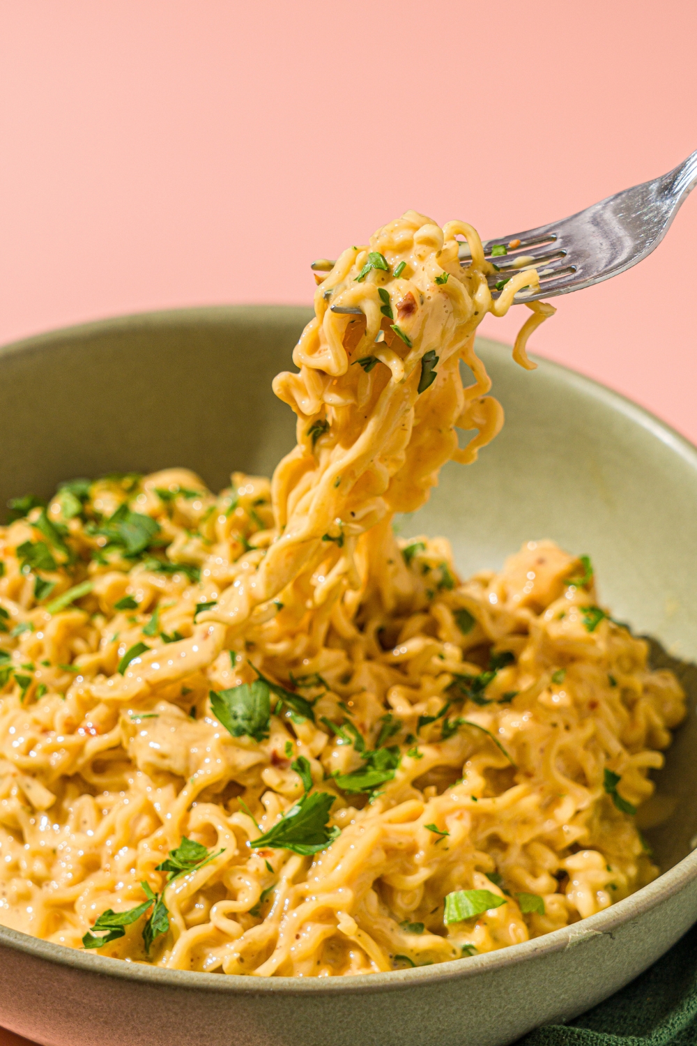 A green bowl with cheesy maggi noodles garnished with fresh cilantro. There is a fork taking a bite of noodles from the bowl. The bowl is on a pink counter.