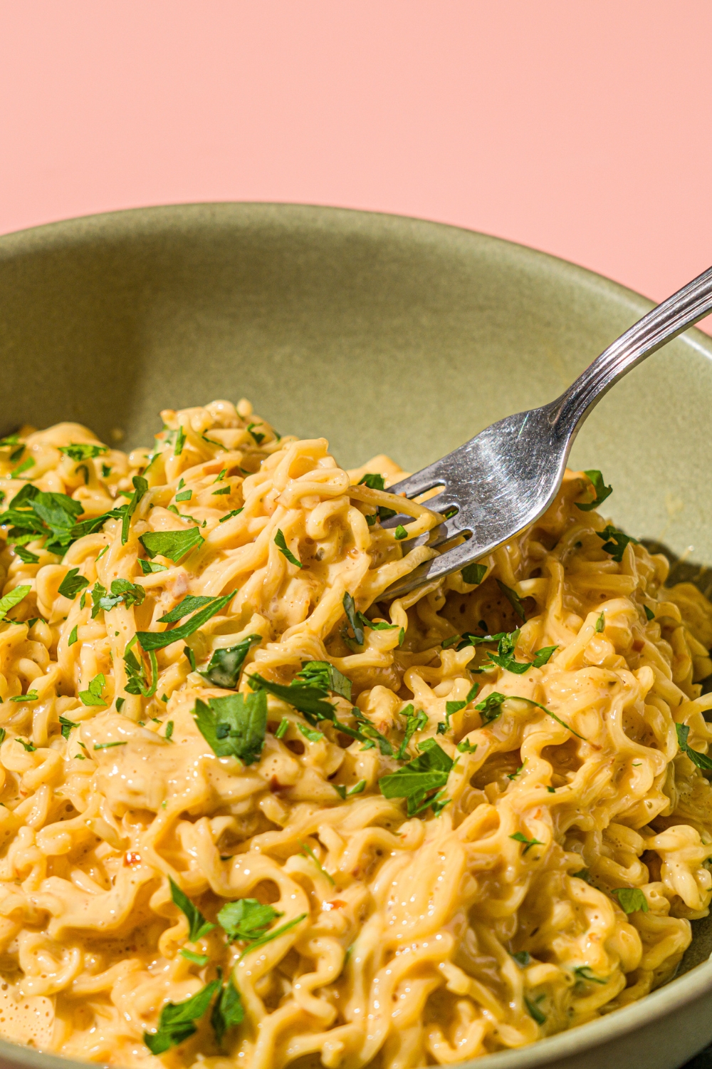 A green bowl with cheesy maggi noodles garnished with fresh cilantro. There is a fork taking a bite of noodles from the bowl. The bowl is on a pink counter.