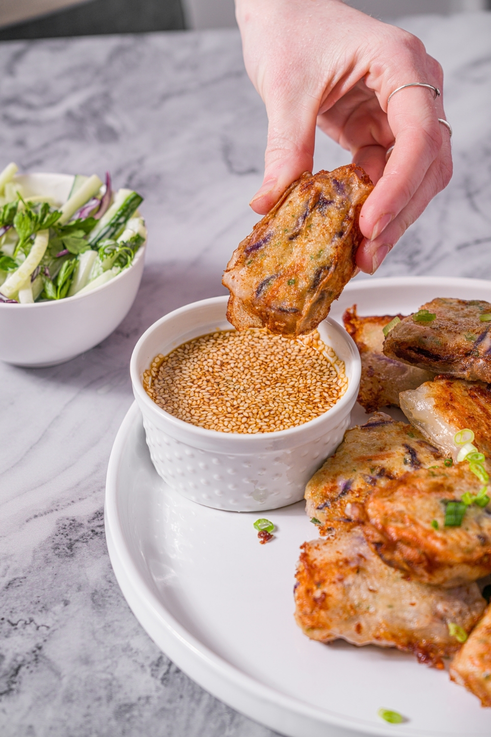 A white plate with chicken and shrimp rice paper dumplings garnished with sliced green onions and served with a small bowl of dipping sauce. A hand is dipping a dumpling into the sauce. The plate is on a marble counter with a small side salad in a bowl.
