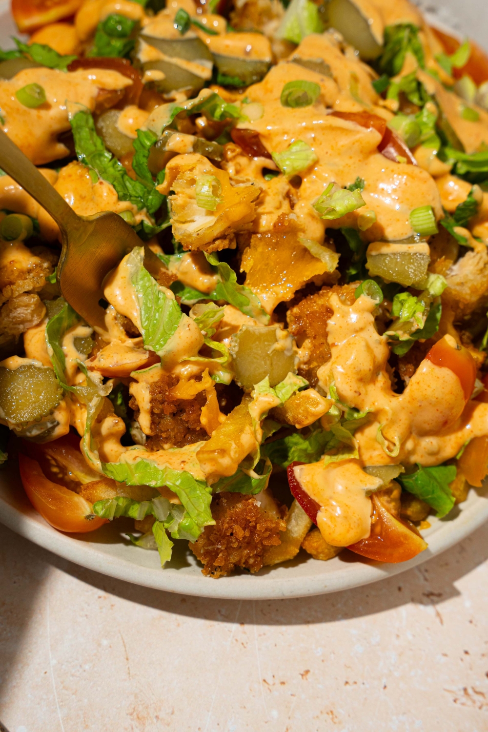 A white plate with chicken burger bowl loaded fries with fries topped with crispy chicken, lettuce, shredded cheese, tomatoes, and pickles drizzled with burger sauce. A fork is taking a bite from the plate. The plate is on a tan counter.