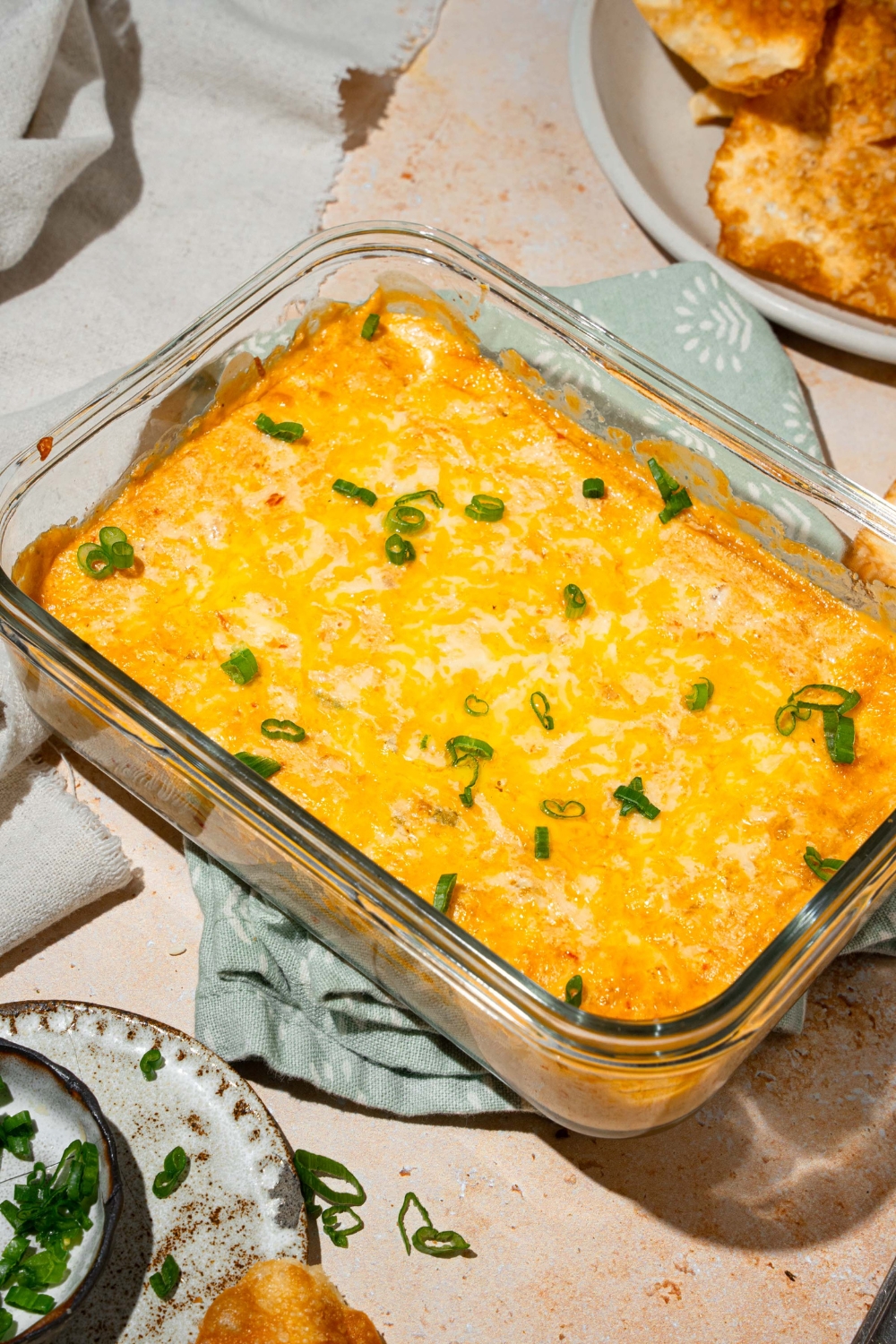 A glass baking dish with baked crab rangoon dip garnished with sliced green onions. The dish is on a tan counter with a bowl of wonton chips, and white cloth napkin.