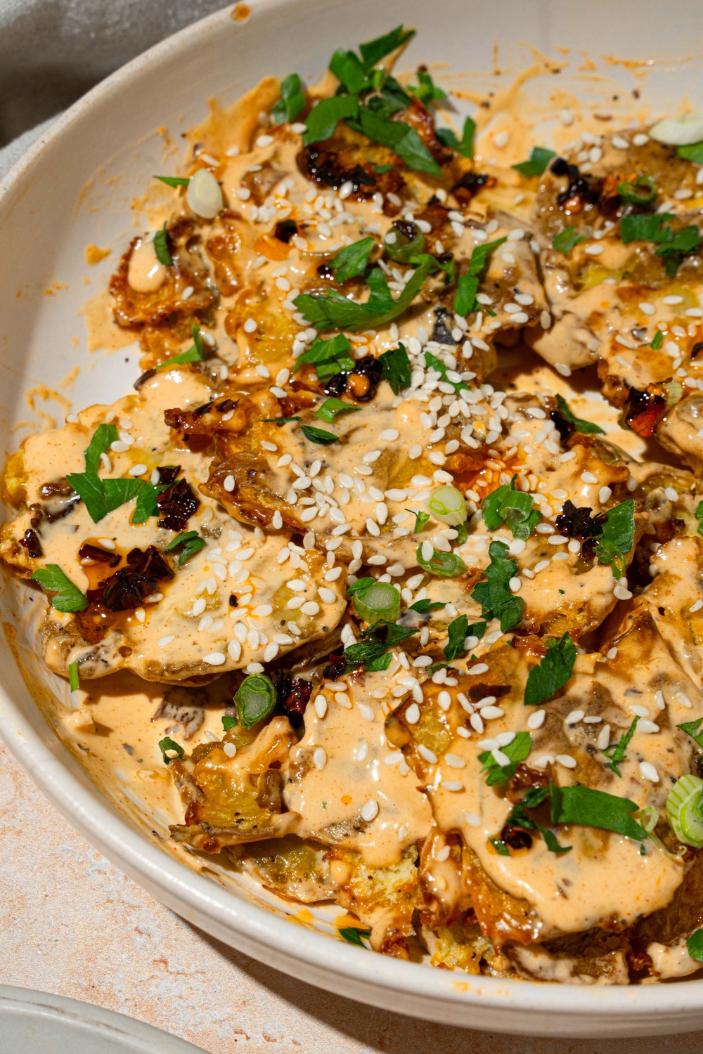 A white bowl with smashed potato salad tossed in chili crisp tahini dressing garnished with sesame seeds and sliced green onion. The bowl is on a tan counter.