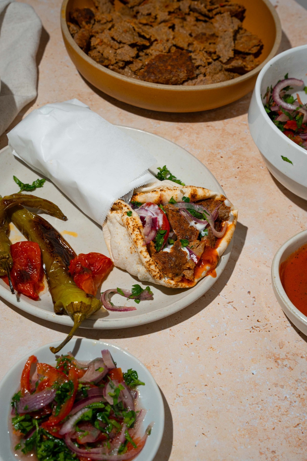 A white plate with a doner kebab wrapped in pita bread with garlic sauce and roasted vegetables with a yogurt sauce. The pita is wrapped in paper. The wrap is on a white plate with roasted green peppers and roasted tomatoes. The plate is on a tan counter.