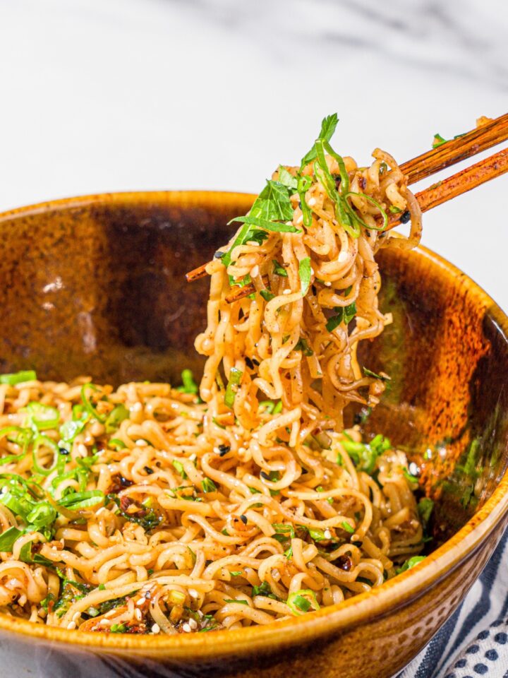 A ceramic bowl with garlic chili oil noodles garnished with sliced green onion and sesame seeds. A pair of chopsticks is taking a bite of noodles from the bowl. The bowl is on a marble counter with a white cloth napkin.