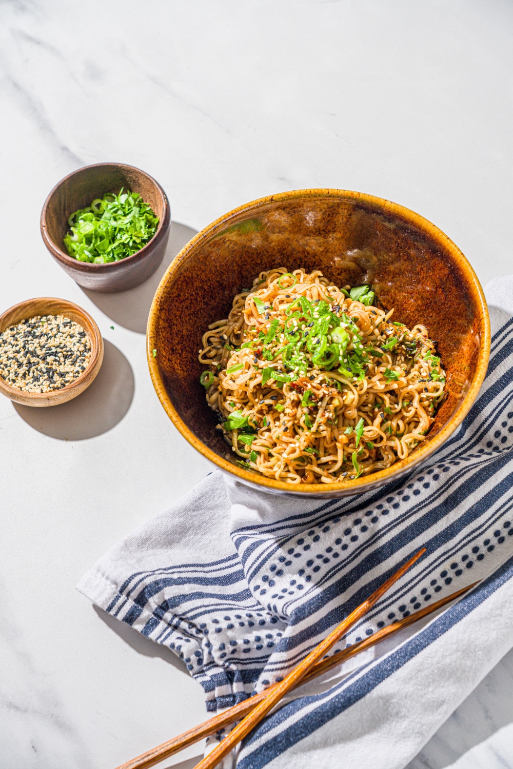 A ceramic bowl with garlic chili oil noodles garnished with sliced green onion and sesame seeds. The bowl is on a marble counter with bowls of garnishes, a pair of chopsticks, and a white cloth napkin.