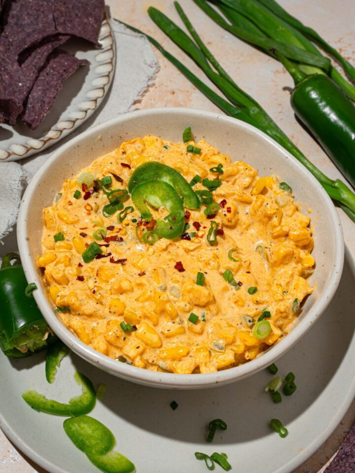 A bowl of jalapeño corn dip garnished with sliced green onion and sliced jalapeños. The bowl is on served on a white plate with sliced jalapeños. The plate is on a tan counter with blue corn chips and green onion.