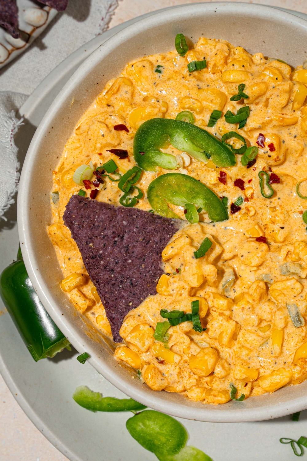 A bowl of jalapeño corn dip garnished with sliced green onion and sliced jalapeños. There is a blue corn chip in the bowl. The bowl is on served on a white plate with sliced jalapeños. The plate is on a tan counter.