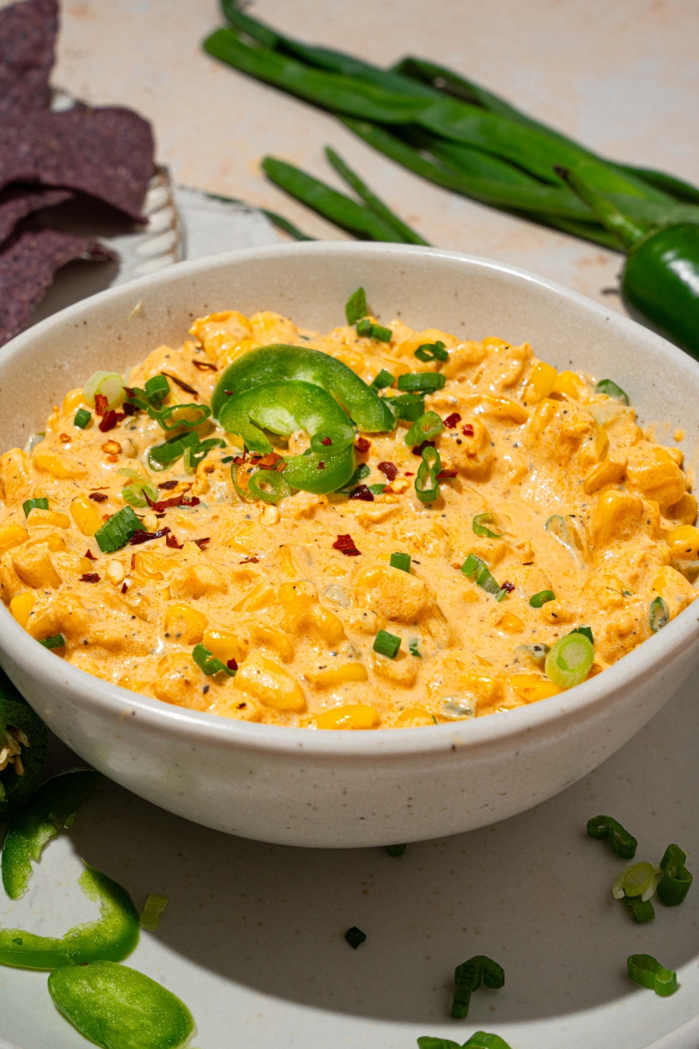 A bowl of jalapeño corn dip garnished with sliced green onion and sliced jalapeños. The bowl is on served on a white plate with sliced jalapeños. The plate is on a tan counter with blue corn chips and sliced jalapeños.