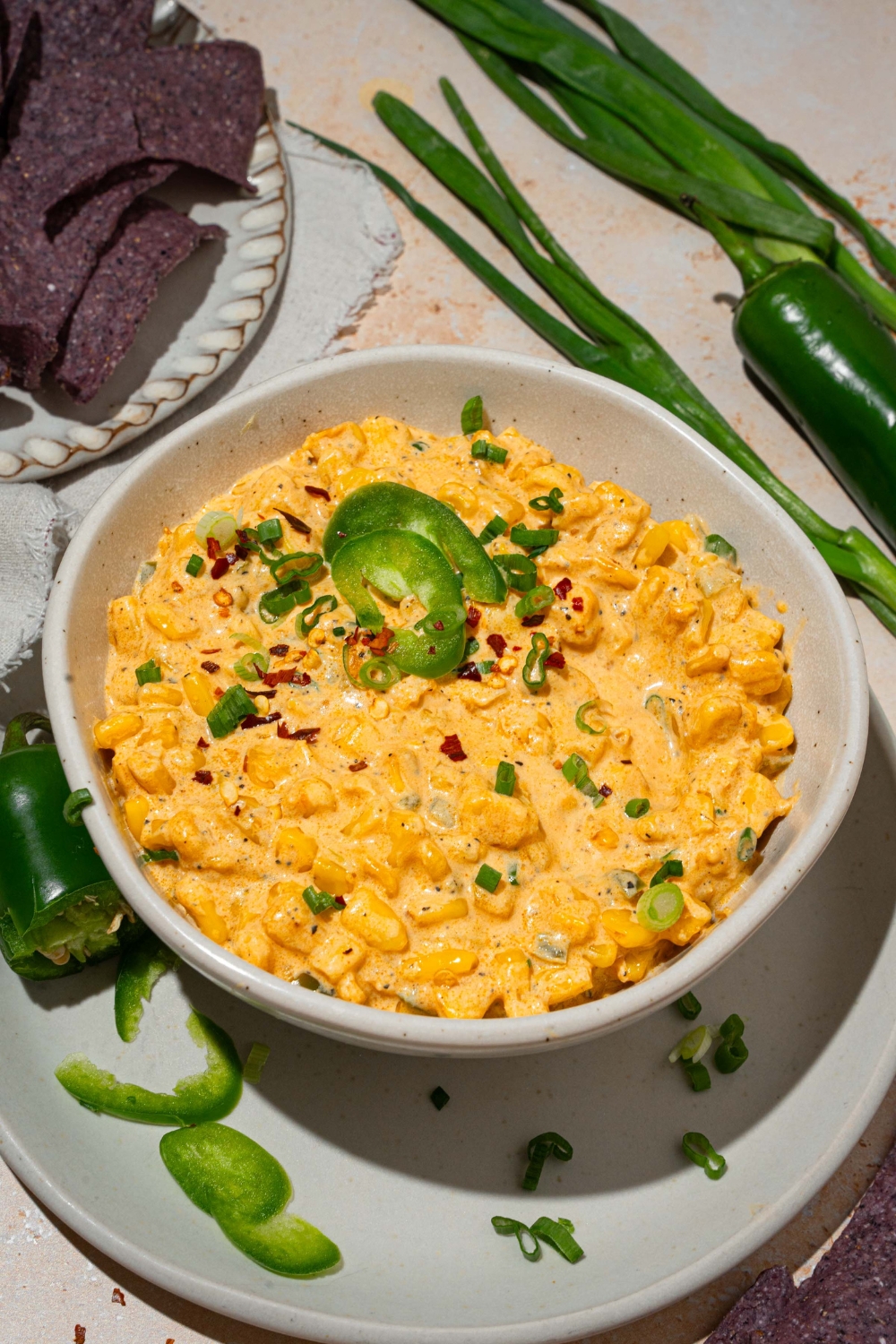 A bowl of jalapeño corn dip garnished with sliced green onion and sliced jalapeños. The bowl is on served on a white plate with sliced jalapeños. The plate is on a tan counter with blue corn chips and green onion.