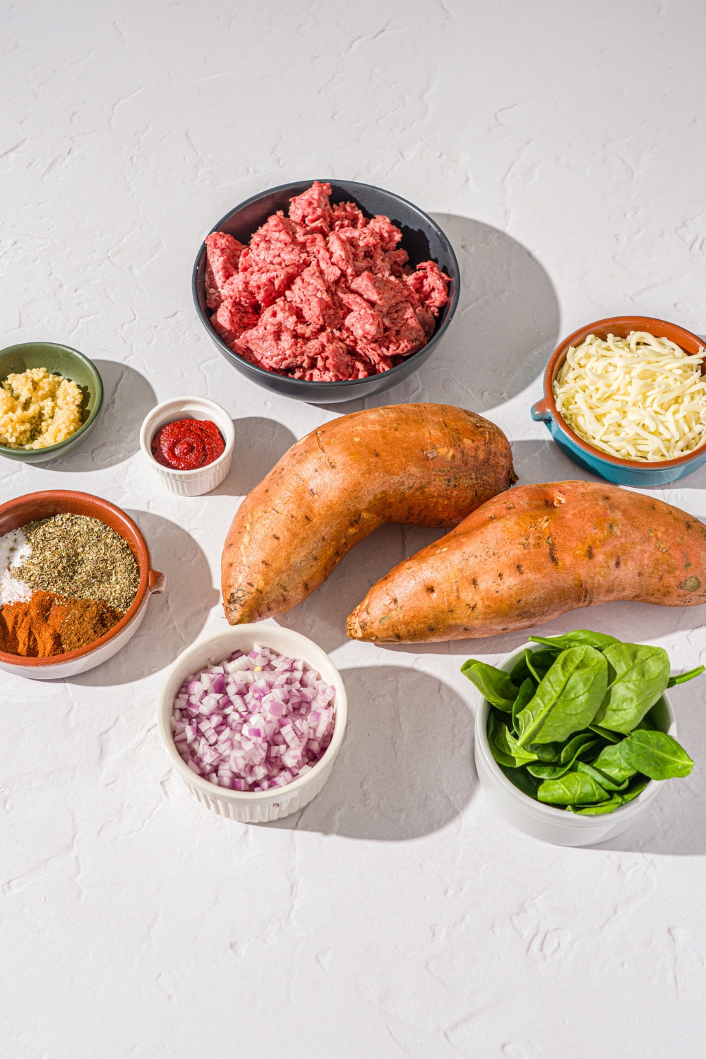 A white counter with several bowls in various sizes containing ingredients to make loaded sweet potatoes including sweet potatoes, ground beef, diced onion, spinach, shredded cheese, garlic, tomato paste, garlic, and seasonings.