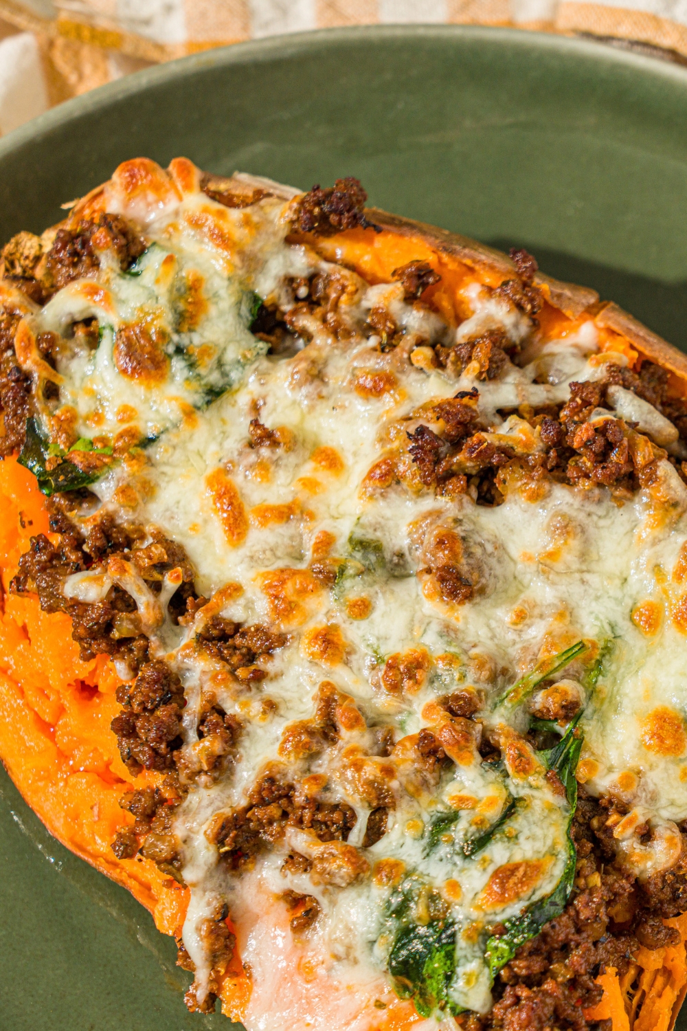 A green plate with a loaded sweet potato sliced open and stuffed with a beef mixture and topped with melted cheese. The plate is on a white counter with a yellow checkered napkin.