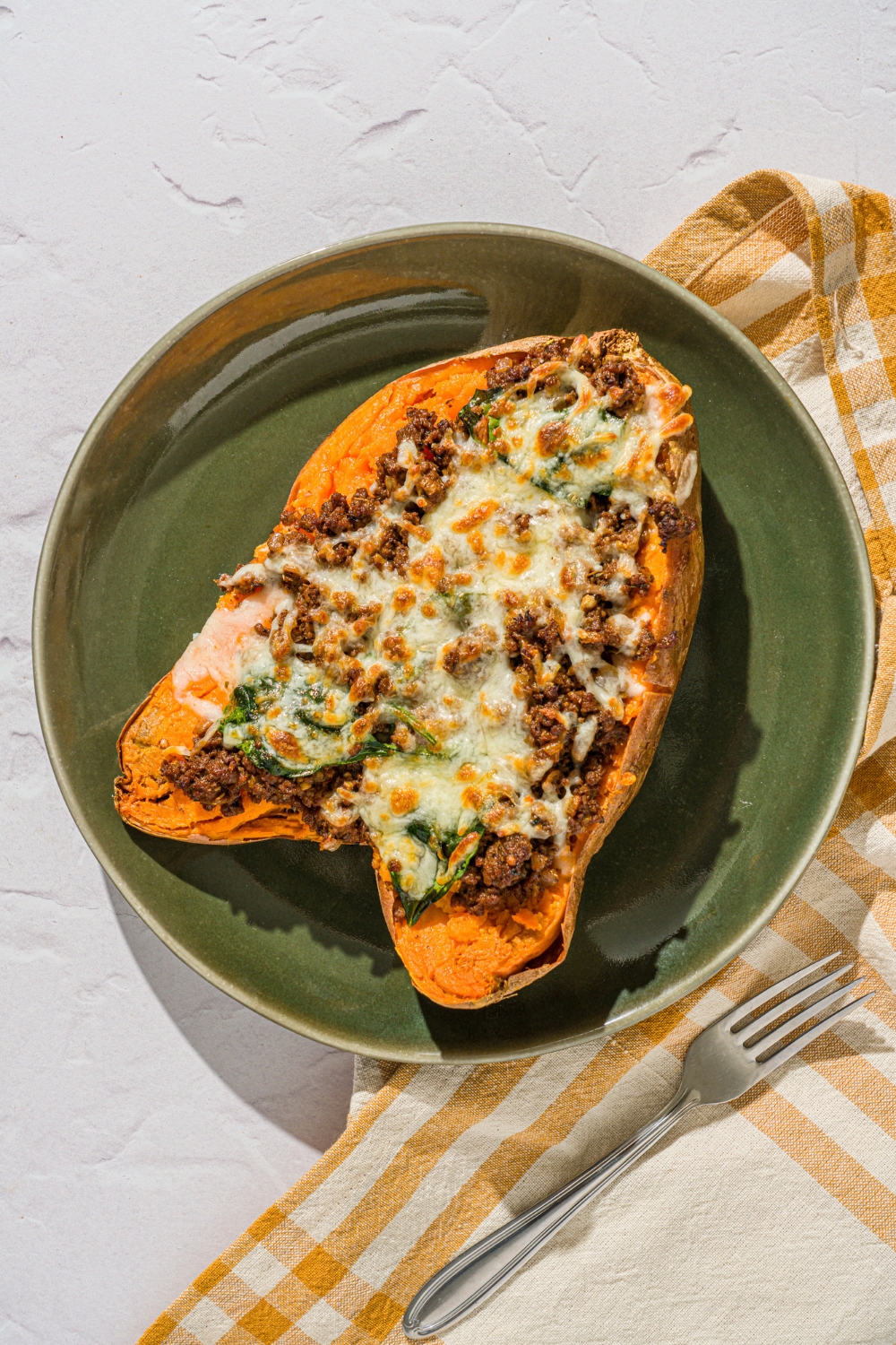 A green plate with a loaded sweet potato sliced open and stuffed with a beef mixture and topped with melted cheese. The plate is on a white counter with a yellow checkered napkin.
