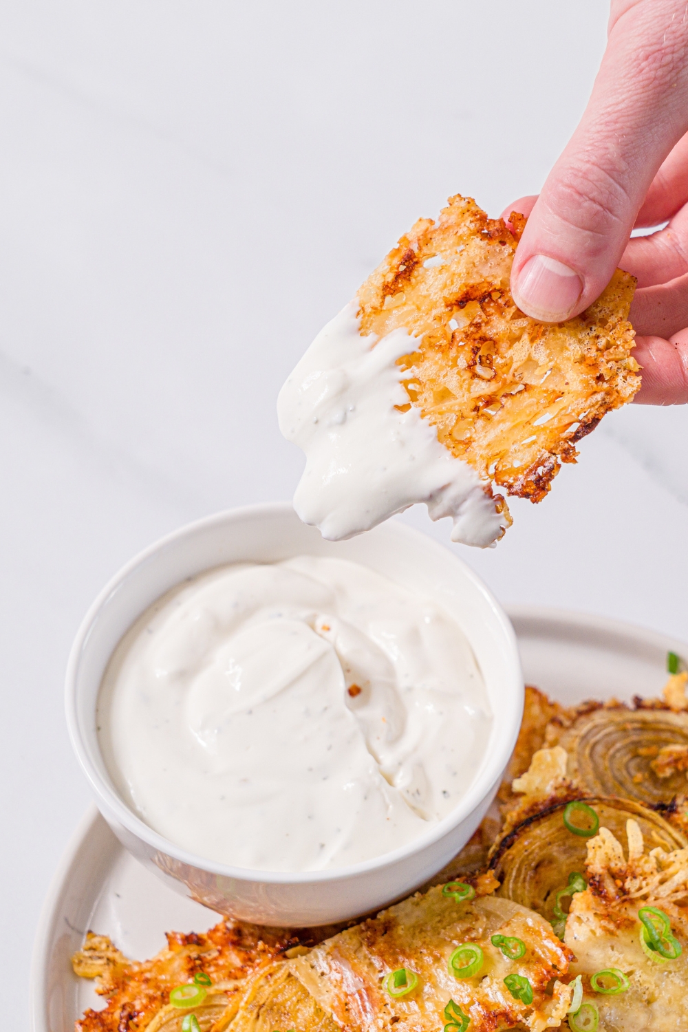 A white plate with baked parmesan onion chips garnished with sliced green onions and small bowl of dip. A chip is being dipped into the bowl. The plate is on a marble counter.