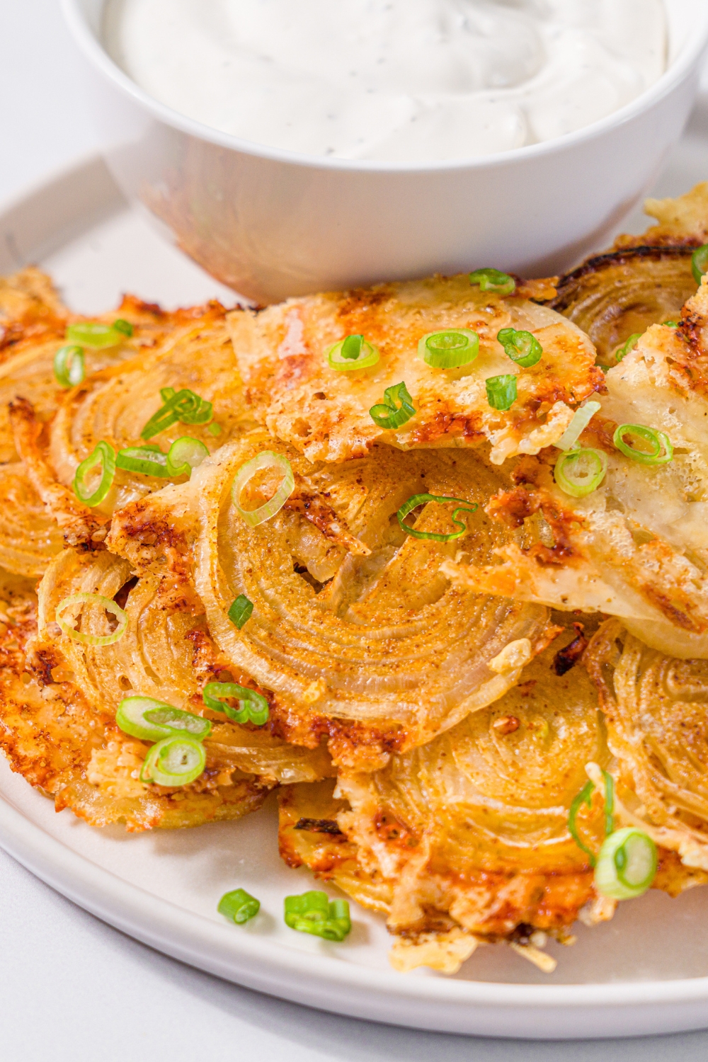 A white plate with baked parmesan onion chips garnished with sliced green onions and small bowl of dip. The plate is on a marble counter.