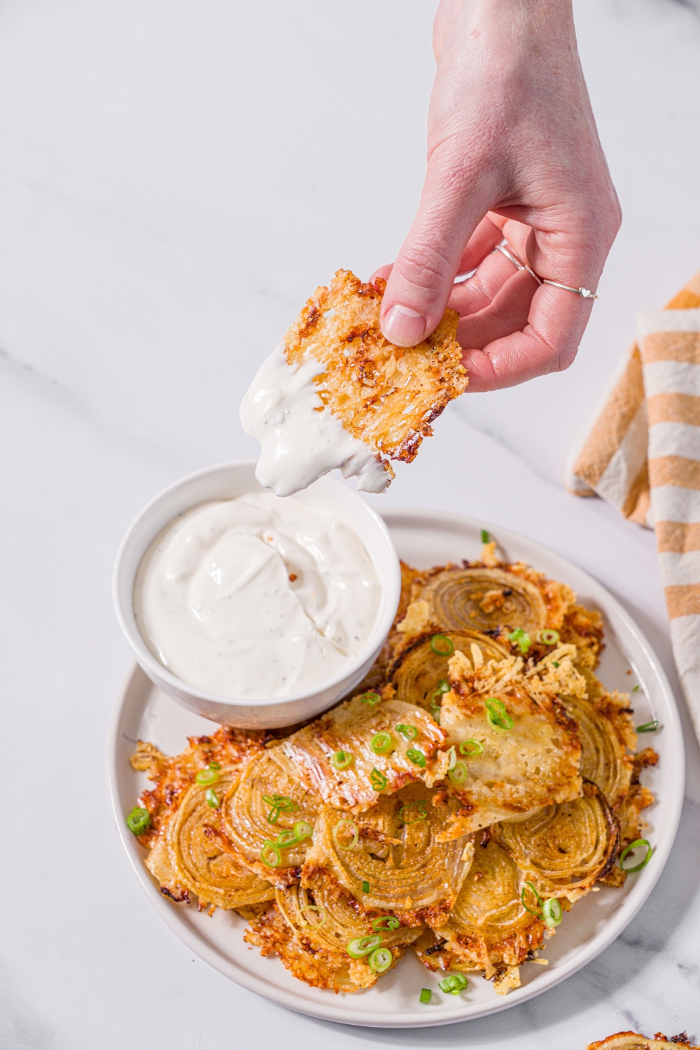 A white plate with baked parmesan onion chips garnished with sliced green onions and small bowl of dip. A hand is dipping a chip into the dip. The plate is on a marble counter with a yellow striped napkin.