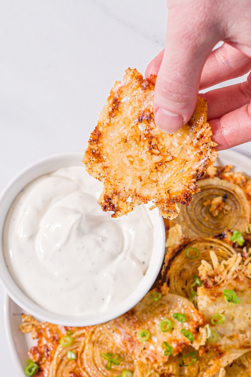A white plate with baked parmesan onion chips garnished with sliced green onions and small bowl of dip. A hand is dipping a chip into the bowl. The plate is on a marble counter with a yellow striped napkin.