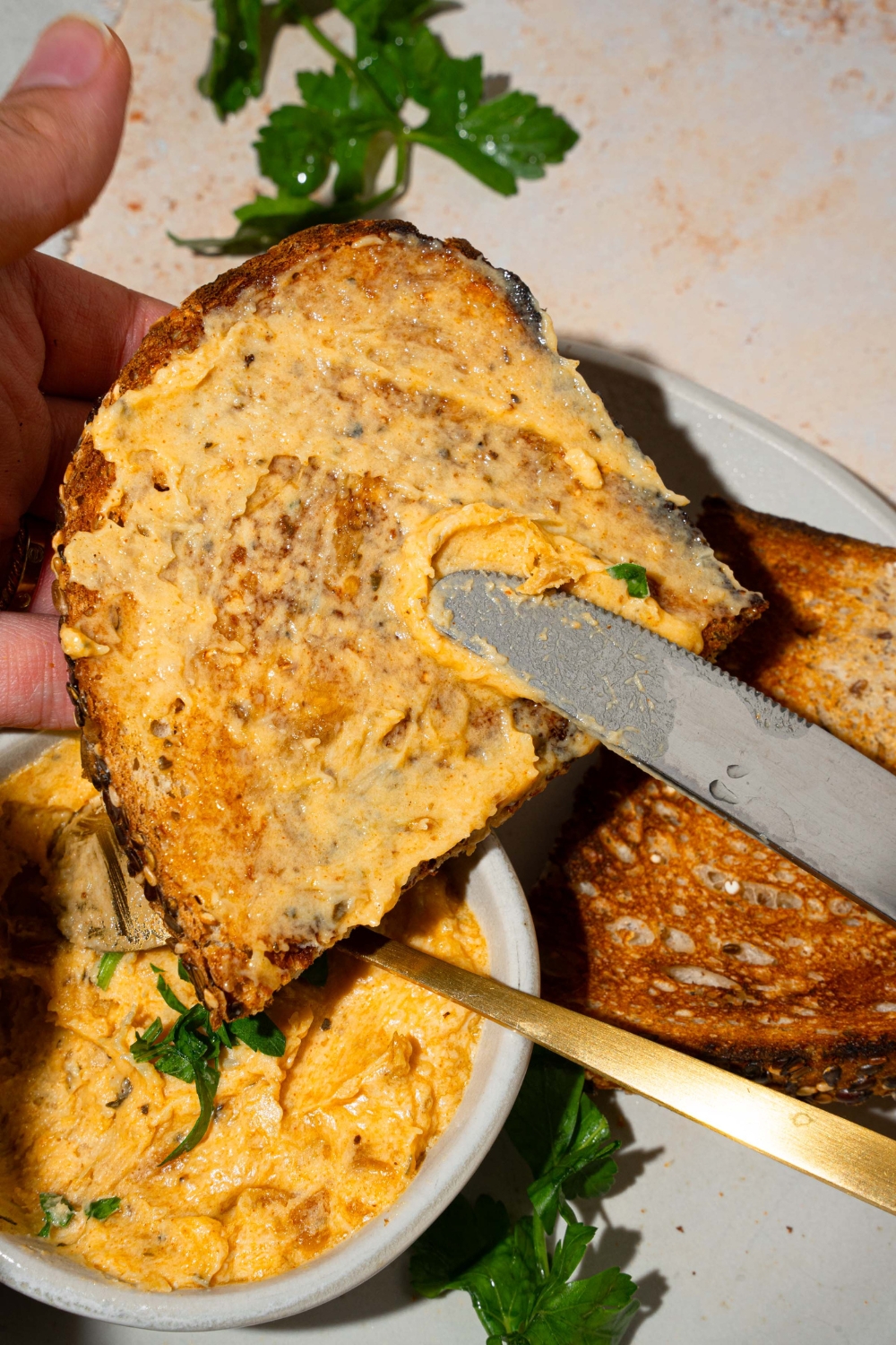 A knife spreading garlic dip over a slice of toasted bread on a white plate with an additional slice of bread and small bowl of garlic dip. The plate is on a tan counter.
