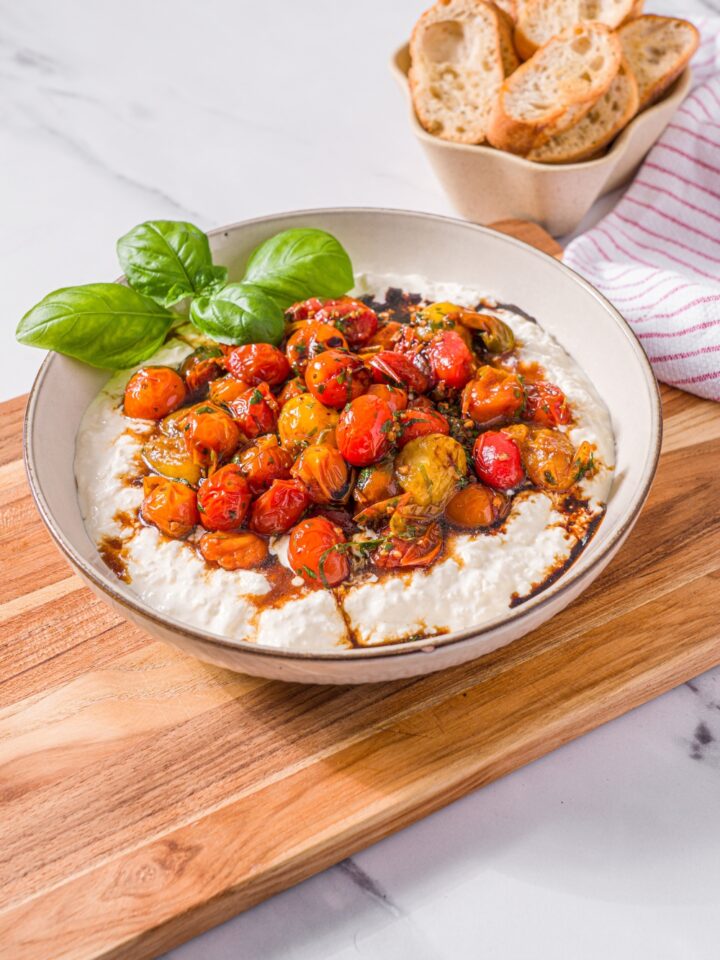 A white bowl with roasted tomato burrata dip garnished with fresh basil. The bowl is on a wooden board on a marble counter with a small bowl of toasted crostini.