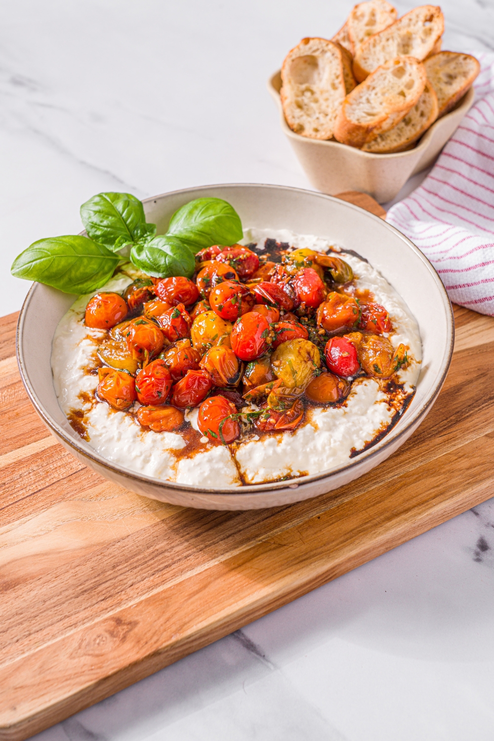 A white bowl with roasted tomato burrata dip garnished with fresh basil. The bowl is on a wooden board on a marble counter with a small bowl of toasted crostini.