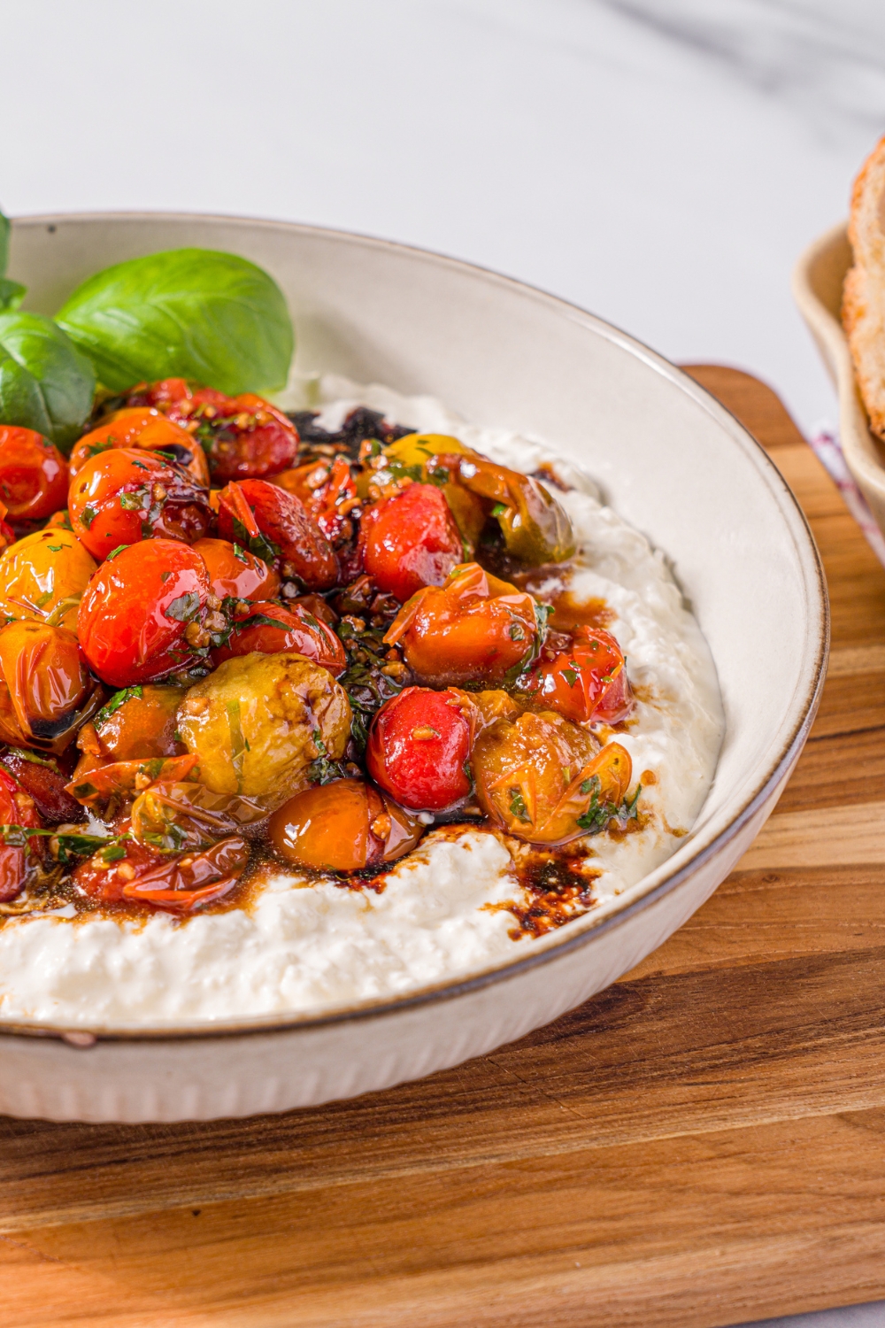 A white bowl with roasted tomato burrata dip garnished with fresh basil. The bowl is on a wooden board on a marble counter with a small bowl of toasted crostini.