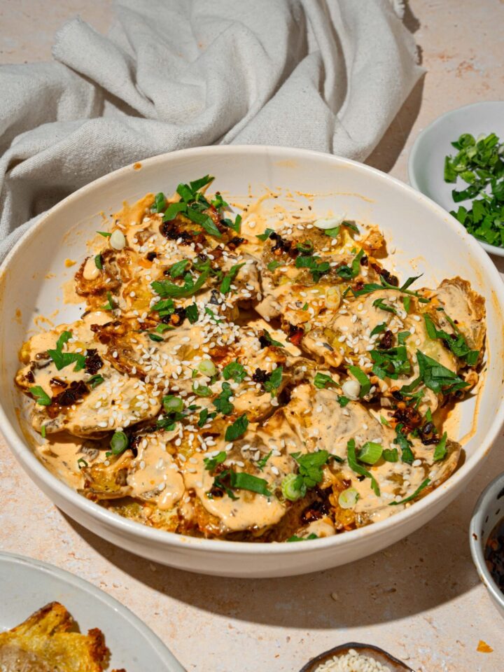 A white bowl with smashed potato salad tossed in chili crisp tahini dressing garnished with sesame seeds and sliced green onion. The bowl is on a tan counter with small bowls of garnishes.