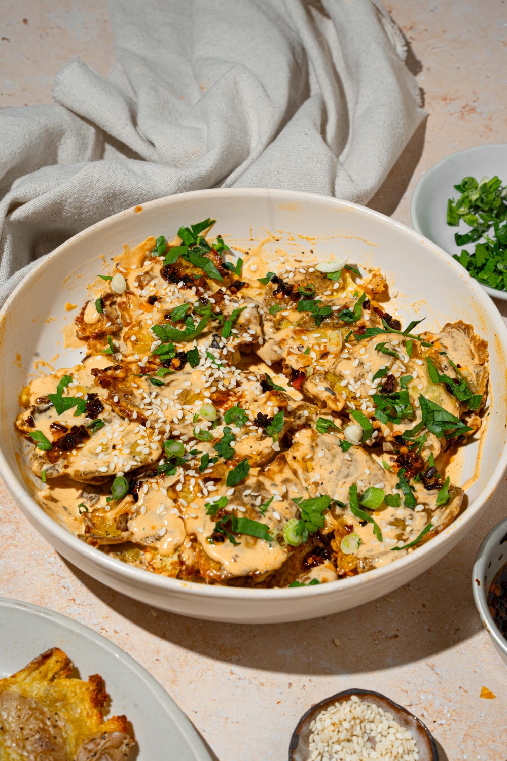 A white bowl with smashed potato salad tossed in chili crisp tahini dressing garnished with sesame seeds and sliced green onion. The bowl is on a tan counter with small bowls of garnishes.