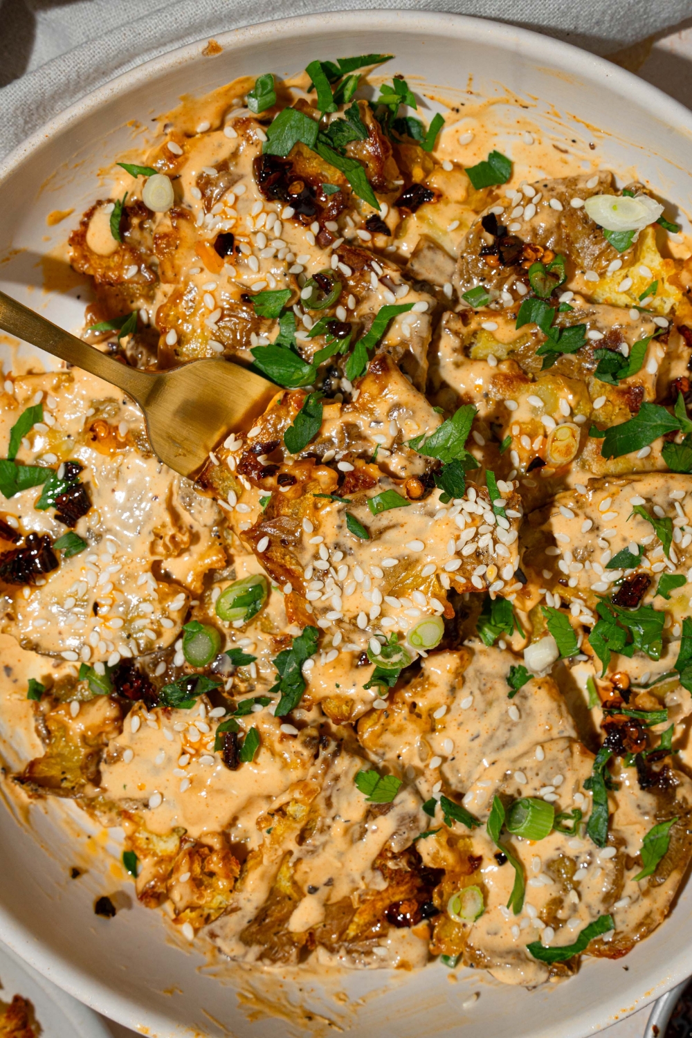 A white bowl with smashed potato salad tossed in chili crisp tahini dressing garnished with sesame seeds and sliced green onion. There is a fork taking a bite of potatoes.
