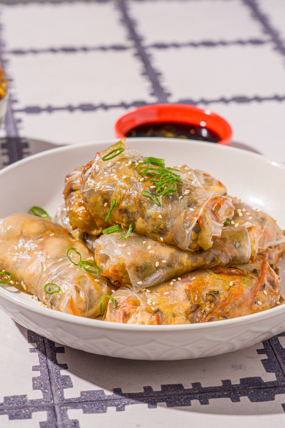 A white bowl with vegetable rice paper dumplings garnished with sliced green onions and sesame seeds. The dumplings are lightly fried. The bowl is on a tiled counter with a pair of chopsticks and small bowl of dipping sauce.