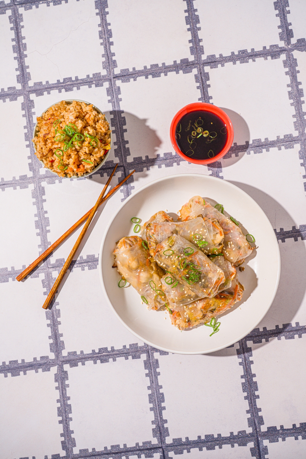 A white bowl with vegetable rice paper dumplings garnished with sliced green onions and sesame seeds. The dumplings are lightly fried. The bowl is on a tiled counter with a pair of chopsticks and small bowl of dipping sauce.