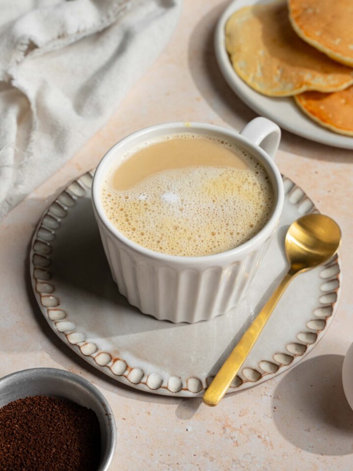 A white mug with Vietnamese egg coffee on a saucer with a spoon. The saucer is on a tan counter with a plate of pancakes and a white cloth napkin.