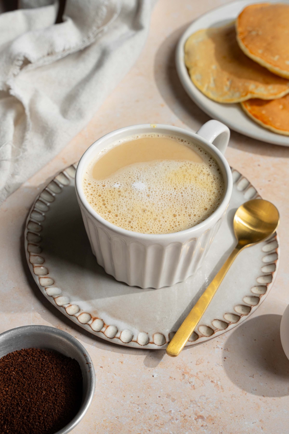 A white mug with Vietnamese egg coffee on a saucer with a spoon. The saucer is on a tan counter with a plate of pancakes and a white cloth napkin.