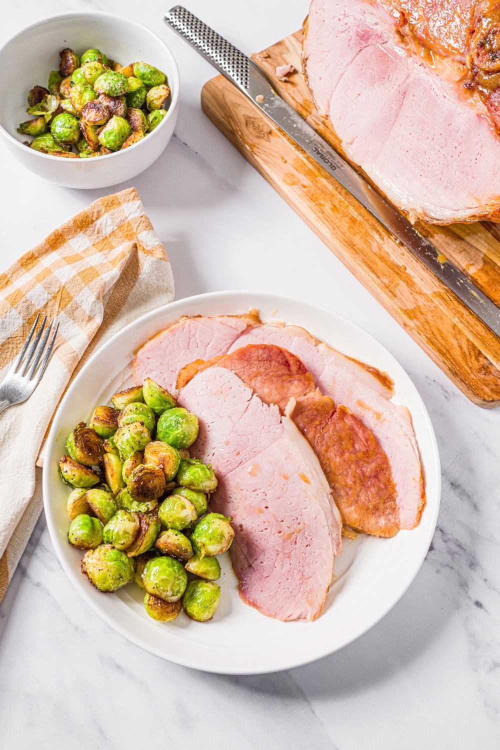 A few few slices of ham shank on a white plate with brussels sprouts. Behind it is a bowl of brussels sprouts and a wooden cutting board with some more ham on it.