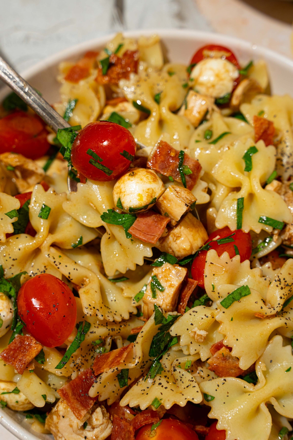 A white bowl of bruschetta chicken pasta with bacon tossed with bowtie pasta, mozzarella cheese, tomatoes, and basil in a balsamic vinaigrette. There is a fork in the bowl.