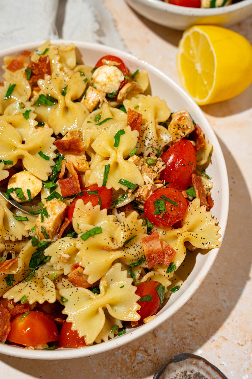 A white bowl of bruschetta chicken pasta with bacon tossed with bowtie pasta, mozzarella cheese, tomatoes, and basil in a balsamic vinaigrette. The bowl is on a tan counter with a lemon and white cloth napkin.