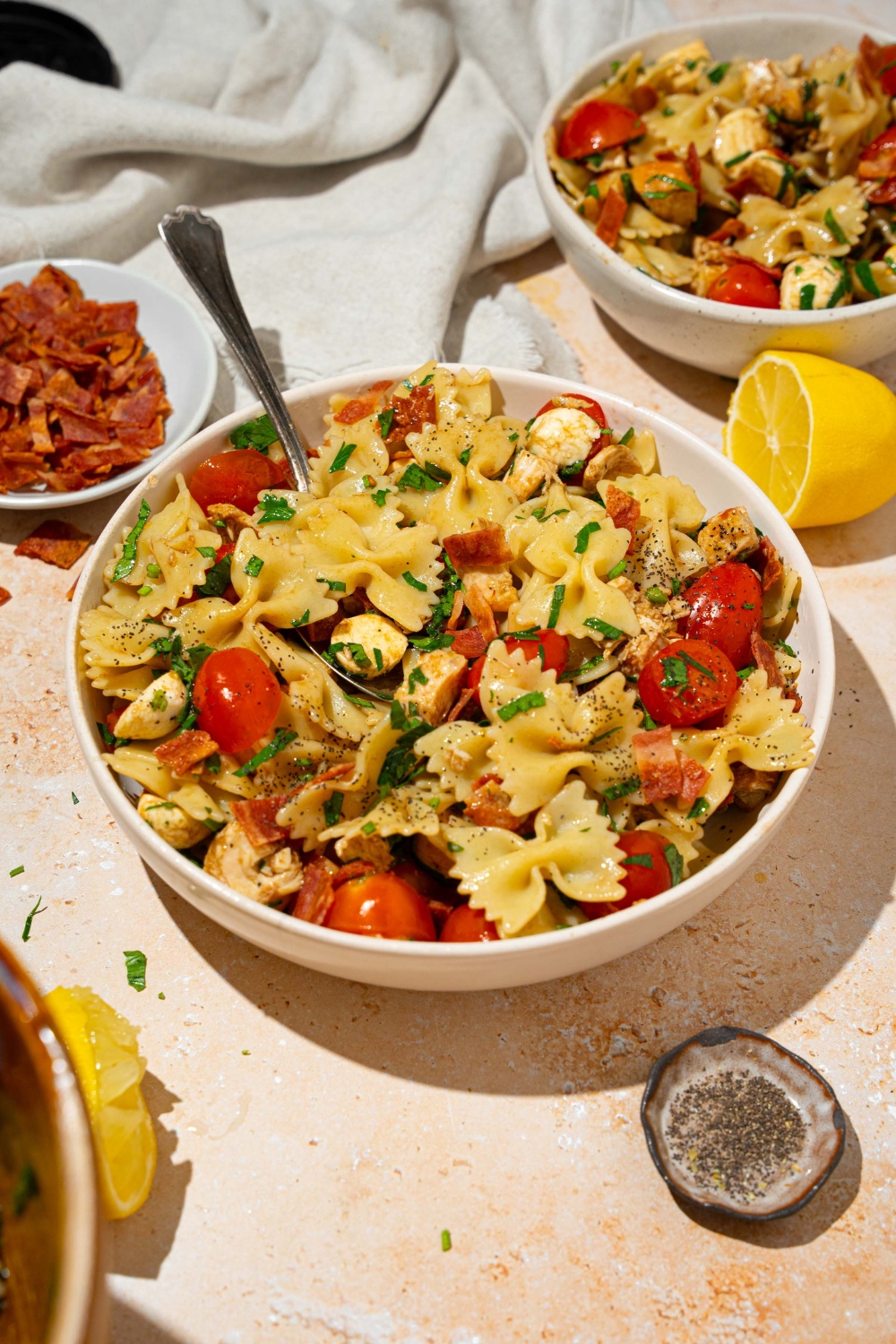 A white bowl of bruschetta chicken pasta with bacon tossed with bowtie pasta, mozzarella cheese, tomatoes, and basil in a balsamic vinaigrette. There is a fork on the bowl. The bowl is on a tan counter with an additional bowl of pasta, small bowl of bacon, and lemon.