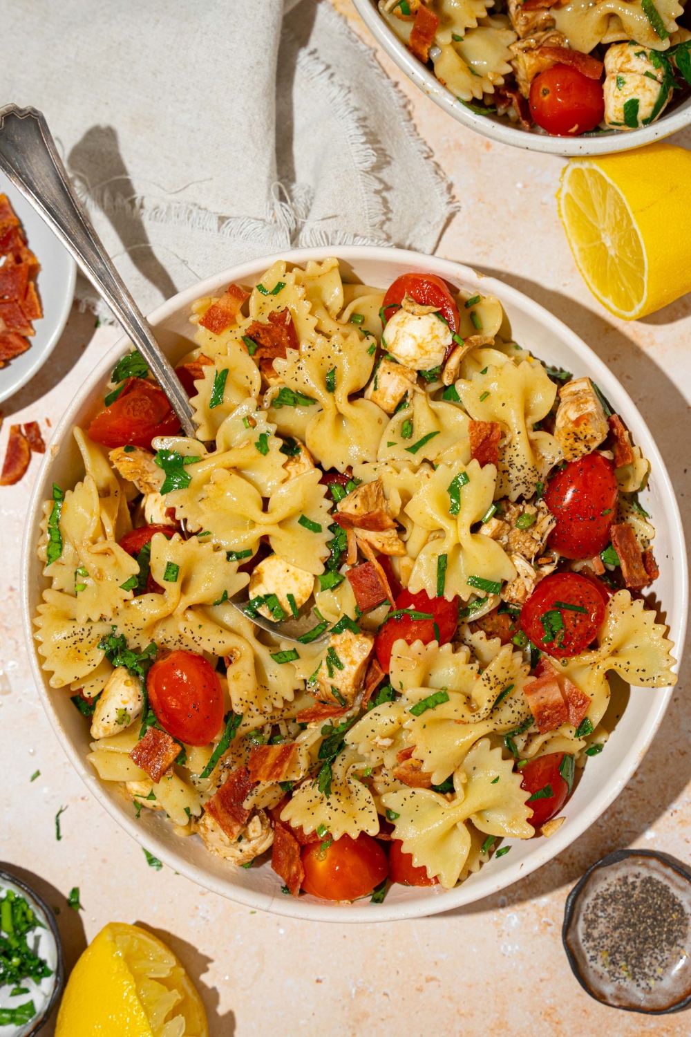 A white bowl of bruschetta chicken pasta with bacon tossed with bowtie pasta, mozzarella cheese, tomatoes, and basil in a balsamic vinaigrette. There is a fork on the bowl. The bowl is on a tan counter with an additional bowl of pasta, small bowl of bacon, and lemon.